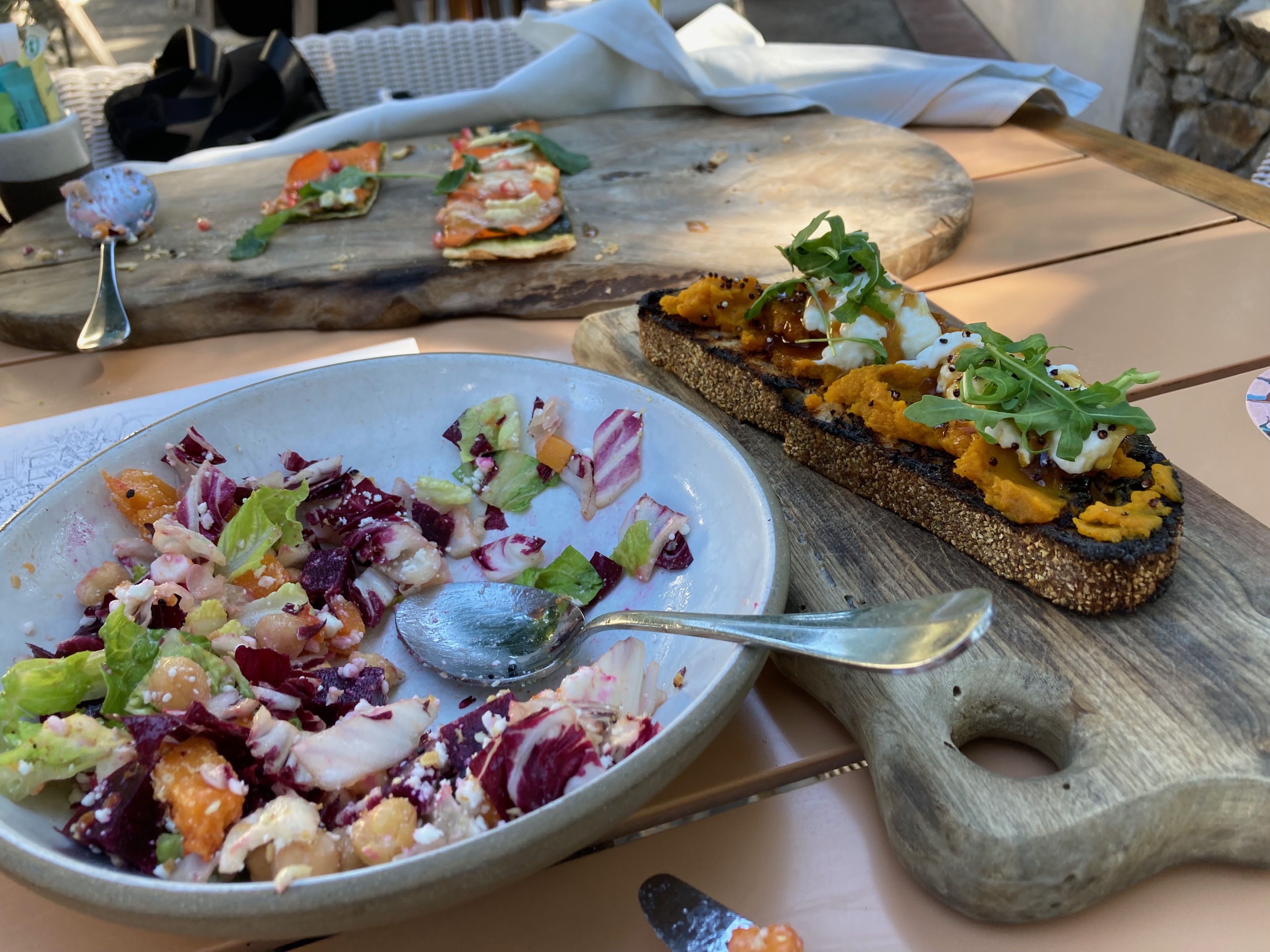 Image of a lovely lunch. A mixed salad in a white bowl. A tartine and a pizza both served on rustic cutting boards. In an outdoor setting with filtered dappled light.